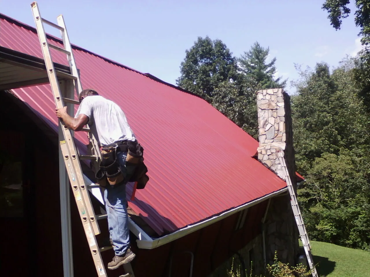 Roofer on a ladder beside a red metal roof and stone chimney — Soffit & Fascia Repair by Roofing Brothers in North Royalton